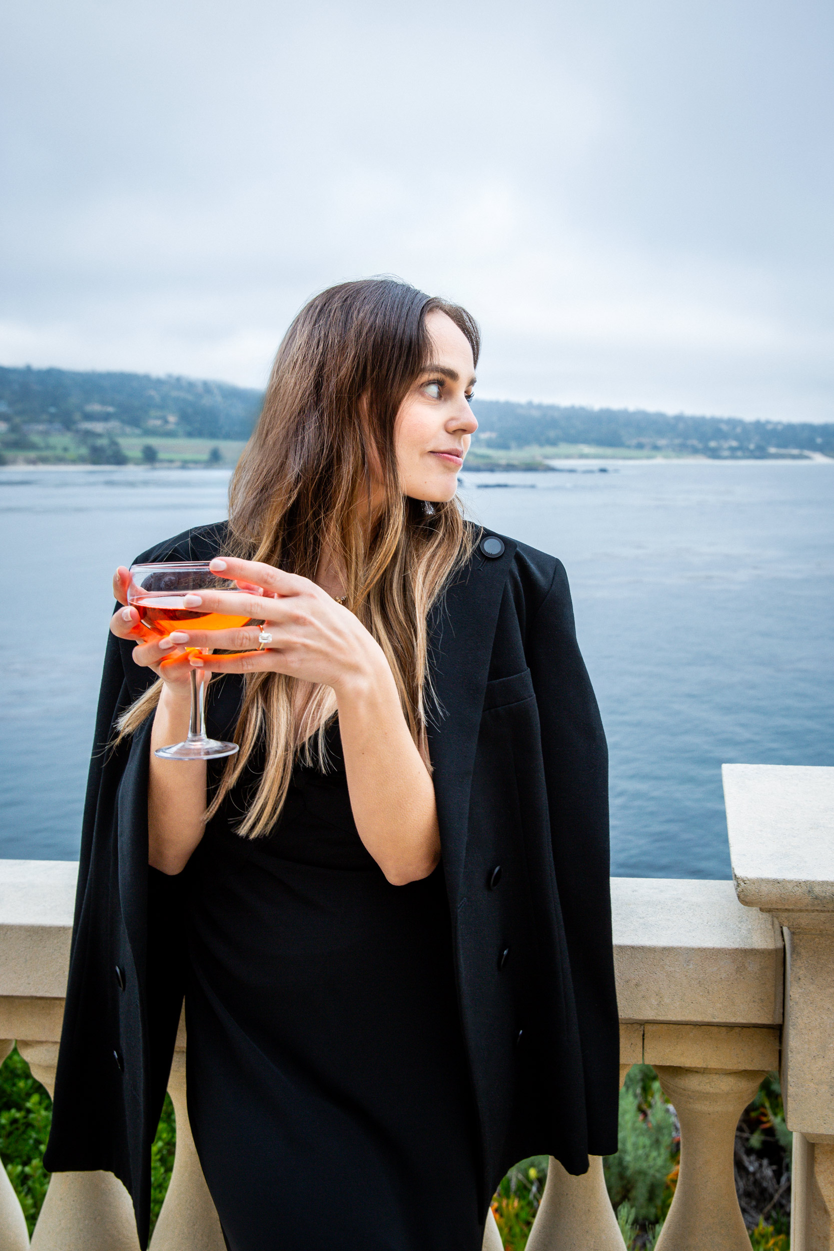 Woman in black blazer holding aperol coupe on stone terrace overlooking Pebble Beach 